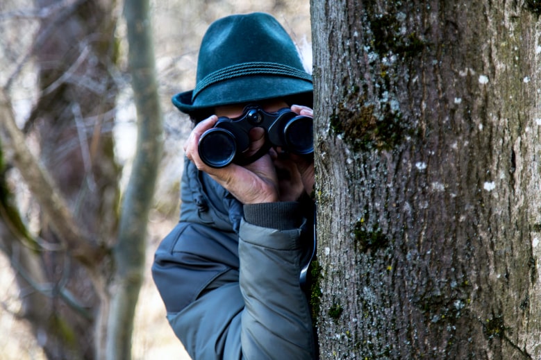 Mann spioniert mit Fernglas hinterm Baum